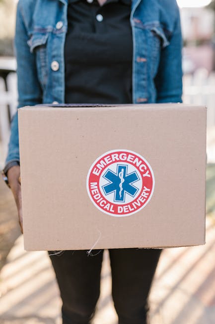A person in a denim jacket and black shirt holding a cardboard box with an emergency medical delivery sticker. The focus is on the box, which is being presented outdoors, with a blurred background of 