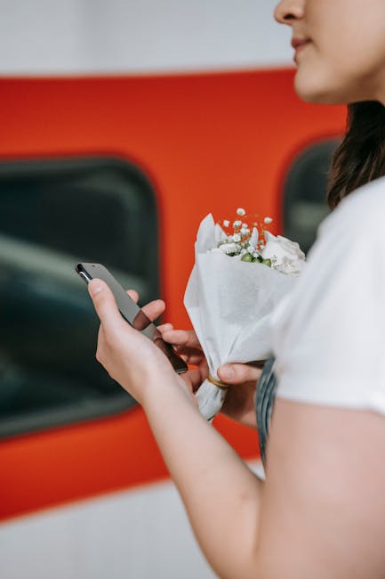 A woman holding a small bouquet of fresh flowers, including white roses and delicate baby's breath, wrapped in white paper with a satin ribbon, standing next to a red vehicle. She is looking at her sm